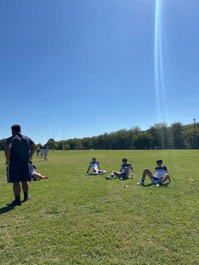 The image shows a group of people on a grassy field under a clear blue sky. A line of trees is visible in the background. In the foreground, a man in a dark shirt and shorts is standing. Further up the field, three people in athletic wear are positioned on the ground, possibly doing exercises. There are additional people near a set of goalposts in the middle distance. The location is San Antonio de Areco, Argentina.