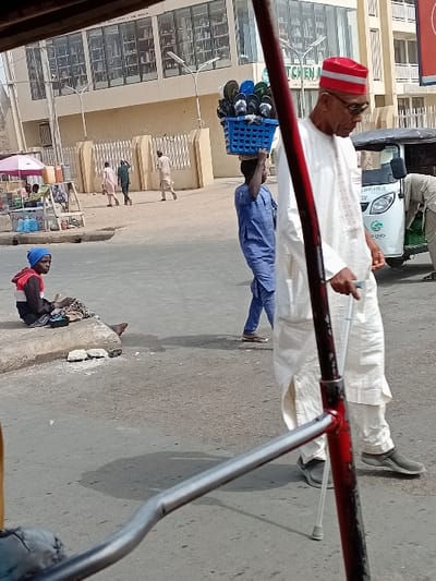 The image presents a daytime street scene in Sani Mai Nagge, Nigeria, viewed from a low perspective, partially obstructed by a red vertical bar and a silver horizontal bar in the foreground.
In the mid-ground, an adult male walks from right to left, wearing a white traditional tunic and trousers, dark grey slip-on shoes, sunglasses, and a red cap with white horizontal stripes. He uses a silver crutch under his left arm, with his right hand in his pocket. Immediately behind him, a younger male, dressed in a blue traditional tunic and trousers, carries a blue plastic basket filled with various colored flip-flops on his head.
To the far left, a young individual with a blue head covering, a red top, and dark patterned pants sits barefoot on a low concrete curb, looking towards the viewer while manipulating items on their lap. To the right, a white and green motorized tricycle is partially visible, with a person standing nearby.
The background features a multi-story building with a light-colored facade and numerous large glass windows. Text on the building includes "SANI MAI NAGGE" and "KITCHEN &". A light-colored, decorative fence runs in front of the building. Several male pedestrians in light-colored traditional garments walk on an unpaved area adjacent to the building. To the left, a small street vendor's stall with a pink umbrella and displayed items is visible. The environment is illuminated by natural daylight, casting shadows.