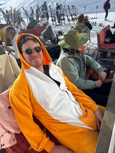 The image captures an outdoor, sunlit scene at a ski resort in St. Christoph, Austria, featuring a bustling social area on a snow-covered mountain. In the foreground, a male subject is seated, wearing a bright orange and white hooded fleece onesie with a white belly panel secured by white buttons. He wears round, reflective sunglasses and looks directly at the camera with a slight smile. To his right, a second individual is partially visible, seated, wearing an olive-green wide-brimmed hat adorned with yellow studs and an olive-green jacket. This individual is actively engaged in handling a dark brown, possibly leather, object with both hands.
The immediate setting includes outdoor seating with red-striped cushions and wooden table surfaces. In the midground, multiple other individuals are seated at similar outdoor tables, some with drinks. Behind these tables, numerous sets of skis and ski poles are stored vertically in metal racks on a snow-covered area. The background reveals a wide, snow-covered mountain slope under a clear sky, with additional skiers and pedestrians visible across the landscape. Shadows are cast across the snow and seating area, indicating bright sunlight.
