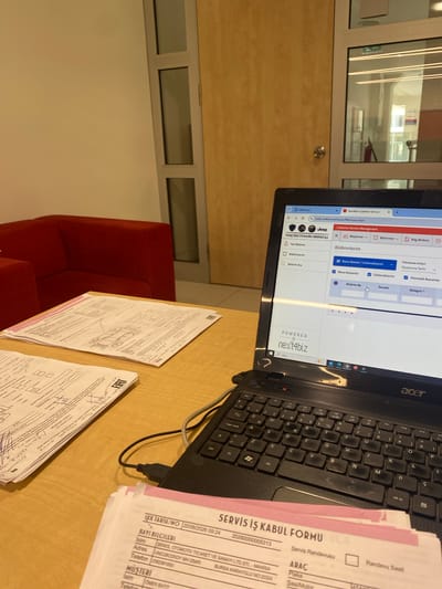 The image shows a close-up view of a desk with a laptop and several papers. The laptop is open and displays a web page from "Next4biz Customer Service" with tabs for "Tofas Türk Otomobil Fabrikası A.Ş.", "Jeep", and "WhatsApp". The laptop screen shows a table with columns for "Bildirim No", "Öncelik", and "Kategori".
In the foreground, there are several sheets of paper. One prominent pink-edged form is partially visible, with the heading "SERVİS İŞ KABUL FORMU" (Service Acceptance Form). Below this, there are fields for "BAKI BİLGİLERİ" (Dealer Information) including "İsim" (Name), "Adres" (Address), and "Telefon" (Phone), and "MÜŞTERİ" (Customer) with similar fields. Another document, also with a pink edge, is visible to the left, with similar fields and handwritten notes. A drawing of an upside-down car is also visible on one of the papers.
The desk appears to be made of wood. In the background, a red sofa is visible to the left. To the right and behind the desk, there is a door with glass panels and a sign that says "EXIT". The overall lighting suggests an indoor environment, possibly an office.
While the image contains documents related to "Tofas Türk Otomobil Fabrikası A.Ş.", and the documents have "MANİSA" and "İZMİR" printed on them, the city of Yunus Emre, Turkey is not directly identifiable from the visual information provided in the image.