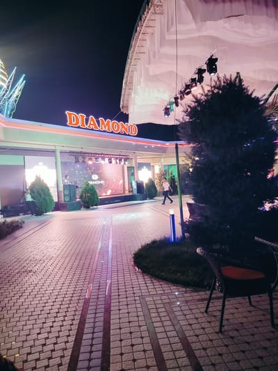 This image depicts an outdoor scene at night, likely at a venue or entertainment establishment in Aksukent, Kazakhstan. The most prominent feature is a large, illuminated sign spelling "DIAMOND" in orange neon letters, situated above an open-air stage. The stage features a large LED screen displaying a graphic with the "DIAMOND" logo, and a person, possibly a DJ, is visible behind a setup of speakers. Several spotlights are suspended above the stage, illuminating the area.
The foreground of the image shows a paved walkway constructed with light-colored square tiles, interspersed with darker, rectangular accent tiles. A dark, evergreen tree dominates the right side of the frame, with its branches obscuring parts of the background. In front of the tree, an outdoor chair with a dark frame and an orange cushion is visible, facing away from the camera.
To the left of the stage, more trees and architectural elements are visible, including a building with a white canopy or awning. The overall atmosphere is festive and brightly lit, characteristic of a night-time public gathering or event. The pavement reflects the colorful lights from the stage and signage, creating a vibrant and dynamic ground-level illumination.