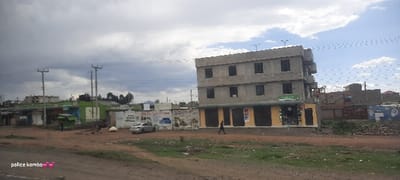 An unfinished three-story building sits on a street corner, its ground floor storefronts closed. A man walks on the sidewalk. Across the street, a row of low, green-roofed buildings stands alongside a painted wall with car advertisements. Power lines stretch across a cloudy sky. The ground is dirt and grassy, with scattered debris. A car is parked near the wall.