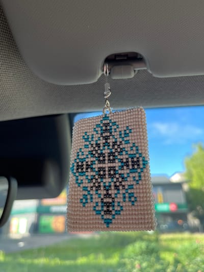A close-up shot shows a beaded car air freshener hanging from the sun visor inside a vehicle. The air freshener is a rectangular piece with a geometric pattern made of white, teal, and black beads. The background is blurred, showing a view through the car's windshield of a sunny day with a blue sky, some trees, and buildings that appear to be in a commercial area. The location is identified as the city of Zhurivka, Ukraine.