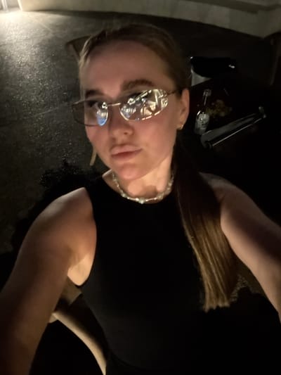 The image shows a woman with blonde hair wearing glasses and a beaded necklace. She is wearing a black top. The setting appears to be a dim or dark location, possibly indoors. There are some objects visible in the background, including what looks like silverware. No information about Jinhua, China can be extracted from this image.