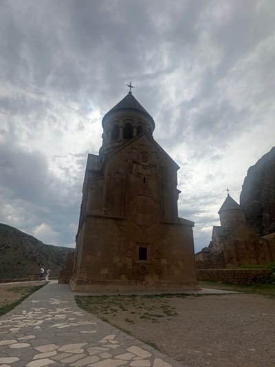 The image displays the Noravank Monastery complex located near the village of Amaghu (as specified, "city of Amaghu") in Vayots Dzor Province, Armenia. The central subject is a large, light-brown stone church, likely Surp Astvatsatsin, featuring a prominent conical dome capped with a Latin cross. The dome rests on an octagonal drum with arched windows. The church's facade exhibits carved stone details, including rosettes and geometric patterns. To the right and behind this main structure, a smaller stone church with a similar conical roof and cross, likely Surp Karapet, is visible, situated against a steep, dark rock face. The surrounding landscape includes arid, vegetated hills on the left and a dramatic rocky cliff on the right. The sky is heavily overcast with grey clouds, showing brighter areas where sunlight penetrates from behind the main church, creating a backlit effect. A paved pathway of irregular flagstones leads towards the central church from the lower left. The ground consists of dry earth, sparse grass, and gravel. Two individuals are present on the left, standing near a low wall; one wears a light shirt and dark trousers, and the other wears a long, light-colored robe, both observing the scene.