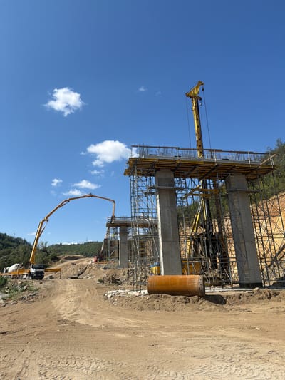 In Kresna, Bulgaria, a large-scale construction project is underway, focusing on the development of a new overpass. The scene is dominated by towering concrete pillars, intricately laced with scaffolding and formwork, indicating the advanced stage of construction. A concrete mixer truck, a common sight at such sites, is positioned on the left, ready to supply the essential material for the project. Adjacent to it, a concrete pump truck extends its articulated arm, poised to deliver concrete to the elevated sections of the overpass. In the background, a dense forest line encircles the construction zone, suggesting a rural or semi-rural setting. The sky above is a clear blue, punctuated by a few fluffy white clouds, implying fair weather conditions conducive to construction work.