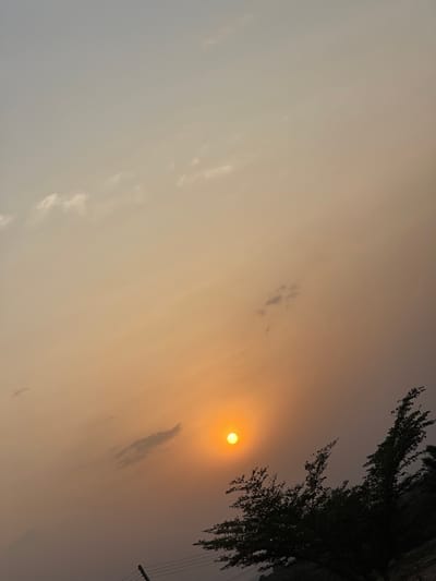 The image captures an outdoor scene in Udi, Nigeria, under atmospheric conditions consistent with sunrise or sunset. The sky occupies the majority of the frame, displaying a gradient color transition from a light grey-blue at the top to a prominent orange and yellow hue around the sun, which then shifts to a hazy, darker grey-brown tone towards the lower right. A distinct, bright orange-yellow solar disc is positioned in the lower-middle left quadrant. Sparse, wispy white and grey clouds are distributed across the upper and middle sections of the sky. In the lower right quadrant, dark silhouettes of tree foliage and branches are visible. The lower-left foreground displays dark, horizontal lines consistent with utility wires, connected to a partially visible vertical pole on the far left edge. No people, specific actions, or interactions are discernible. The environment appears open, indicative of a rural or semi-rural landscape.