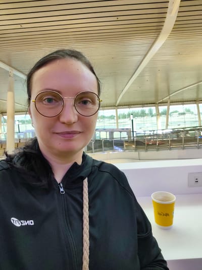 A woman with dark hair and glasses is taking a selfie in what appears to be an airport or a large, modern public space with a high, slatted ceiling. She is wearing a black hooded jacket with a white logo on the left chest and a braided rope strap is visible. To her right, on a white surface, sits a yellow McCafe disposable cup. In the background, through large windows, an outdoor area with greenery and possibly parking lots is visible. The overall environment suggests a travel hub. The specific location in Tambon Mai Khao, Thailand is not identifiable from the image itself.
