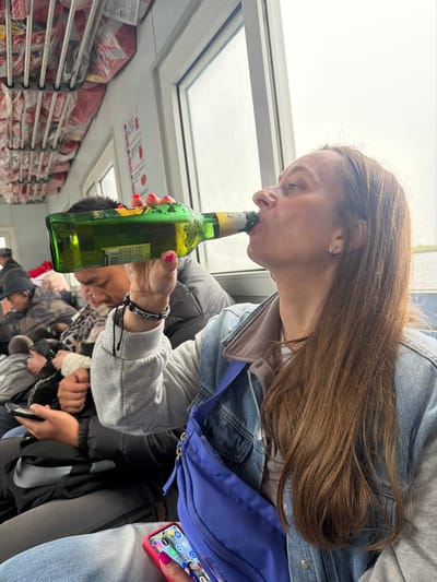 An overhead, slightly tilted perspective captures a young woman drinking from a green glass bottle within what appears to be the interior of a transport vessel or large boat, possibly a ferry, in Leninskoe, Russia. The woman, with long brown hair, wears a denim jacket over a gray hoodie and has a white wireless earbud in her left ear. Her left hand, adorned with pink nail polish, holds the bottle to her mouth. The bottle features a label with "330ml" and a partial barcode ("69299..."), along with some indiscernible Asian script.
To her right, a bright blue fanny pack or sling bag hangs, and her right hand holds a smartphone in a red case, displaying app icons and small profile pictures on its screen. She also has a patterned bracelet on her wrist. In the background, other passengers are visible but out of focus, suggesting a crowded environment.
The setting includes white-framed windows or skylights above, through which a bright, overcast sky is visible, indicating daytime. Along the left side of the cabin, rows of red-wrapped packages, possibly cargo or emergency supplies, are neatly stacked on metal racks. A partially visible red sign with white text is also present above these racks. The lighting is natural and bright from the windows. The overall scene suggests travel or transport, with the focus on the woman's casual activity of drinking.