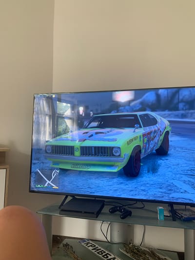 The media file shows a television screen displaying a video game. The game appears to be a racing game set in a possibly snowy or icy environment, with a bright green race car with racing decals as the central subject. The car has the brand name "DECLASSE" visible on its side. In the background of the game, there are hints of buildings and trees.
Beside the television, a glass shelf is visible, holding a PlayStation controller, a game case, and some other small objects. To the right of the shelf, a dried plant arrangement is placed, and a license plate with Ukrainian text ("КРИВБА") is attached to the wall.
The scene is indoors, with the television as the focal point. The lighting suggests it could be daytime, given the reflections on the screen, but the game's environment depicts a cooler, possibly wintery atmosphere. No people are clearly depicted, other than a partial view of a person's thumb in the bottom right corner. The overall mood is casual, centered around gaming.