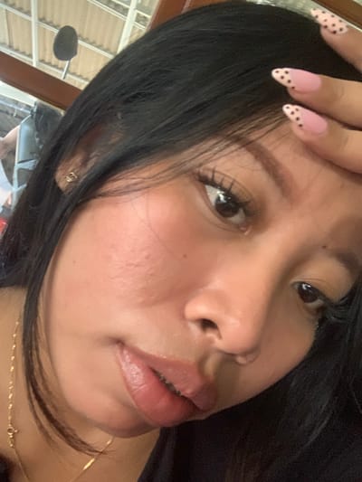 The image is a close-up, overhead shot of a young woman's face. She has dark hair, dark eyes, and light brown skin. Her left hand is resting on her forehead, and her fingernails are painted pink with black polka dots. She is wearing a gold necklace. The background is blurred but appears to be an indoor setting with a ceiling structure. The woman is looking down and slightly to the right. There is no discernible location in Jajag, Indonesia, visible in the image.