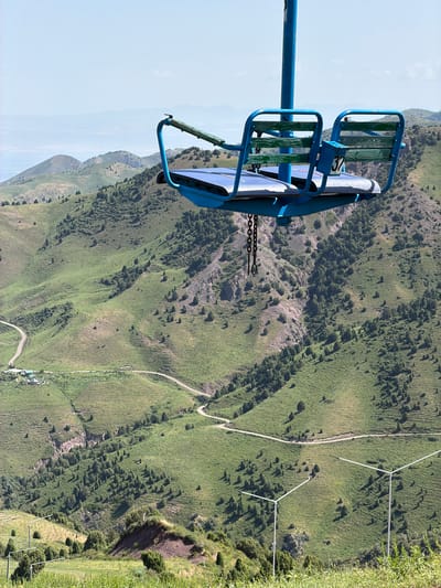 This image, taken in the vicinity of Kashka Suu, Kyrgyzstan, features a ski lift chair in the foreground, suspended against a backdrop of lush, green, mountainous terrain. The chair, painted blue, has two seats with green wooden slats. A metal chain hangs below the chair. In the distance, winding dirt roads are visible, along with scattered trees and some buildings, suggesting a rural or mountainous resort area. The sky is a pale blue, and the overall impression is one of a serene, natural landscape.