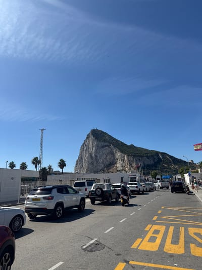 The image shows a sunny day in a border crossing area, likely between Gibraltar and Spain. A prominent rock formation, resembling the Rock of Gibraltar, dominates the background. Several vehicles are parked along the roadside, including cars, a motorcycle, and what appears to be a van or bus. Yellow markings on the asphalt indicate a bus lane. In the distance, a Spanish flag is visible flying from a pole. Palm trees add to the Mediterranean feel of the scene.
On the right side of the image, large yellow lettering on the road clearly spells out "BUS". There is also a sign on a building that reads "ADUANA", which translates to "Customs" in Spanish, further confirming the location as a border crossing. The sky is a clear, bright blue, suggesting fair weather. The time of day appears to be daytime, given the strong sunlight and clear visibility. The overall impression is a busy but orderly scene at an international border.