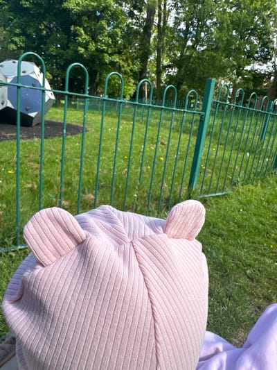 A close-up shot shows a section of a green metal fence in a grassy area, likely a park. Behind the fence is a tree and a black and white geometric structure. In the foreground is part of a pink ribbed hat, possibly with animal ears. The image appears to be taken at ground level, and judging by the information given (Macclesfield, United Kingdom), it's potentially within a public park or recreational area.