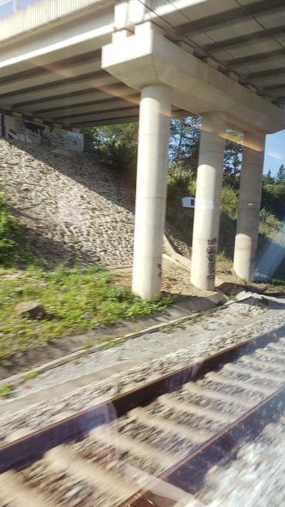 An elevated concrete overpass, supported by thick cylindrical pillars, crosses over a set of train tracks in Horný Hričov, Slovakia. The pillars are marked with spray-painted graffiti. The ground beneath the overpass is covered with loose stones, and a grassy embankment with scattered small flowers runs alongside the tracks. Trees are visible in the background, and the sky is a clear blue. The image appears to have been taken from a moving vehicle, as evidenced by the motion blur on the train tracks and surrounding environment.