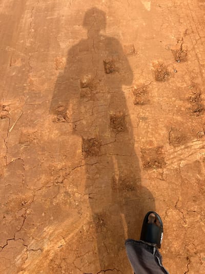 The scene depicts a cracked, reddish-brown dirt ground illuminated by direct sunlight, casting a distinct human shadow. The shadow of an upright individual, likely male, extends from the top-center towards the bottom of the frame. In the bottom-right corner, the lower portion of a human leg is visible, clad in faded denim trousers with a frayed hem, and wearing a dark slide sandal featuring a white logo. The ground exhibits numerous irregular cracks indicative of dry conditions. Multiple roughly square or rectangular depressions are patterned across the dirt, some containing looser soil, possibly imprints from heavy equipment or objects. Faint tire tracks are present on the right side of the ground. A small, light-colored twig and scattered pebbles are visible on the surface. The image is taken in Ilishan, Nigeria.