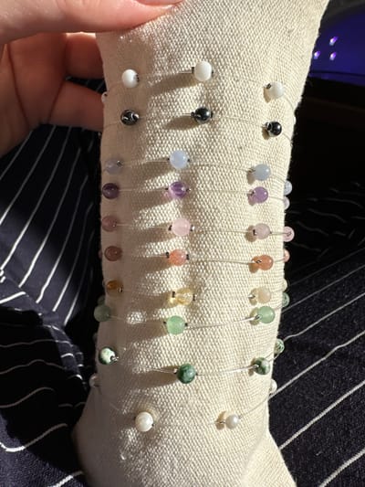 The image shows a hand holding a padded roll covered in off-white fabric. Strung on fine, transparent cords and held in place on the roll are a number of beads of various colors and shades including white, black, blue, purple, pink, orange, yellow, and green. The background consists of a dark blue and white striped fabric. The location in the city of Galiche, Bulgaria is not discernible from the image.