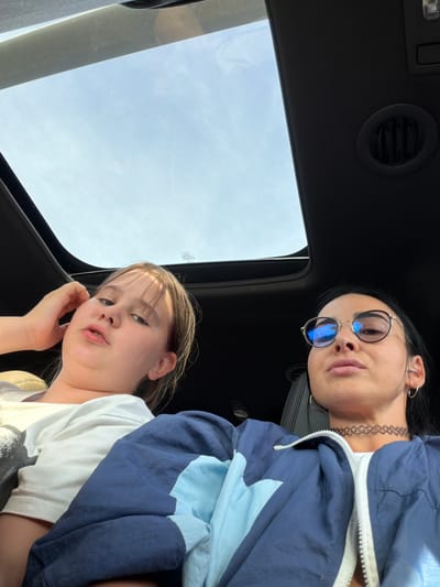 Two young females are inside a vehicle, looking up at the camera through the sunroof. The subject in the foreground is a girl with blonde hair and blue eyes, wearing a white t-shirt. She has her right hand raised to her cheek, and her mouth is slightly open. Behind her, a young woman with dark hair wears round sunglasses and a choker necklace. She is looking down at the camera and is dressed in a blue and light blue jacket. The scene appears to be outdoors during the day, with a clear sky visible through the sunroof. The interior of the car is dark, with a vent visible on the ceiling. The image is a selfie taken from a low angle.