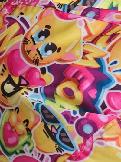 A close-up image displays a section of a brightly colored textile, located in Donchevo, Bulgaria. The fabric features a repeating cartoon pattern characterized by stylized yellow animal faces, possibly cats, each rendered with large light blue eyes, black pupils, pink cheeks, and thin black whiskers. These figures are outlined in black and white. The design incorporates an assortment of bold, graffiti-style elements including letters (e.g., a yellow and pink 'M'), hearts, and abstract shapes. The predominant colors are yellow, fuchsia pink, deep purple, and light blue, with secondary accents of green and black. The fabric exhibits gentle folds, and a horizontal seam is visible near the top of the frame, suggesting it is part of an item of clothing or another manufactured textile. No individuals, explicit actions, or external environmental details are present within the image frame.