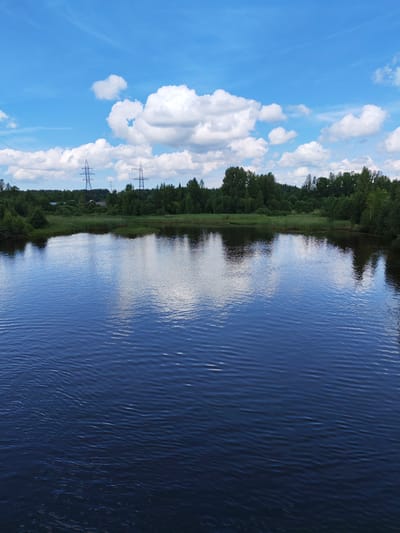 A panoramic, eye-level, full outdoor shot captures a body of water reflecting a blue sky with fluffy white clouds, situated in the village of Akhimovo, Russia. The water, a deep blue, shows gentle ripples on its surface. The far shore is lined with dense green trees and tall reeds, with two tall, dark electricity pylons visible above the tree line, one more prominent than the other. A thin, lighter-colored transmission line stretches between the pylons. In the distance, houses are subtly visible nestled amongst the trees. The lighting suggests a bright, sunny day.