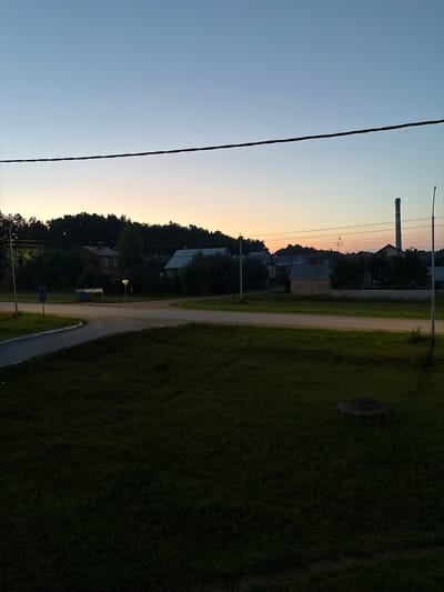 This media file shows a rural scene in Birsk, Russia, at dusk. The sky transitions from a clear blue to a soft orange and yellow hue near the horizon, indicating the setting sun. A thin, dark power line stretches diagonally across the frame from the lower left corner, suggesting the presence of electricity infrastructure in the area.
The landscape features a dirt road winding through a grassy area. On one side of the road, several houses and buildings are visible, nestled amongst dark green trees. Some of these structures have pitched roofs, typical of residential buildings. A tall industrial chimney stands prominently in the background, hinting at nearby industrial activity. A few streetlights are also present, adding to the twilight ambiance.
On the other side of the road, the terrain is covered in dense, dark green grass, with a circular object that appears to be a manhole cover or grate embedded in it. The overall atmosphere is peaceful and quiet, typical of a rural evening. There is no visible text in the image.