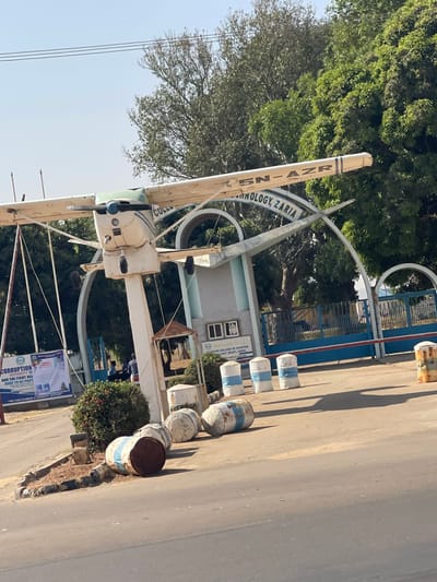 The image displays the main entrance of the Nigerian College of Aviation Technology (NCAT) in Basawa, Zaria, Nigeria, under clear daylight conditions. A white, single-engine propeller aircraft, bearing the registration 5N-AZR, is mounted on a tall white pole, serving as a prominent static display monument. Behind this monument, the institution's main gate features light blue architectural arches and pillars, encompassing a central building with windows and signage that reads "NIGERIAN COLLEGE OF AVIATION TECHNOLOGY (NCAT), ZARIA" and "WELCOME TO". A blue metal gate is visible beyond the pillars. An asphalt road occupies the foreground, leading towards the entrance. To the left of the road, a landscaped island holds green bushes, a small rusted metal structure, and several discarded, heavily rusted metal barrels, some painted with faded blue and white stripes. White and blue striped bollards line the paved entrance path. A large white banner on the left side of the entrance displays partial text indicating "CORRUPTION" and "THE FIGHT AGAINST". In the background, near the gate, indistinct figures are visible, with one individual appearing to be on a motorcycle. Green trees with dense foliage are present behind the main entrance structure.