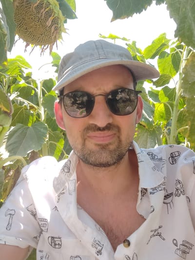 A man wearing a light gray baseball cap, dark sunglasses, and a white short-sleeved shirt with black line drawings of various objects such as an axe, a backpack, and a grill stands in front of a sunflower plant. The man is looking at the camera and has a beard and a mustache. The background consists of green leaves and a bright sky, suggesting an outdoor environment. The image appears to be taken in a rural or garden setting. The location is identified as Yoav Regional Council, Israel.