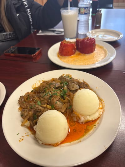 The image shows a table setting in a restaurant, possibly in Des Plaines, United States of America. Two plates of food are visible. The plate on the left contains what appears to be stuffed peppers in a creamy sauce. The plate on the right features two scoops of mashed potatoes, liver and onions, and a reddish-orange sauce or gravy. A glass of milk, a water bottle, and a bottle of condiment are situated next to the plate of stuffed peppers. A Calvin Klein jacket and a cell phone are near the plates. The table has a dark brown surface.