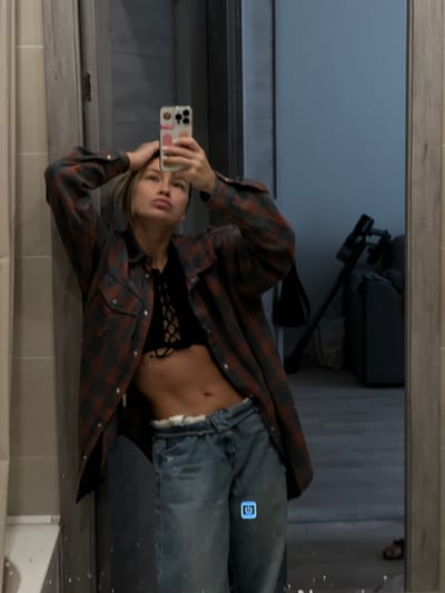 A person, possibly female, is reflected in a bathroom mirror. The person is holding a smartphone, presumably taking a selfie. The person is wearing blue jeans, a black lace top, and a red and gray flannel shirt. Reflected in the background of the mirror are a baby stroller and a blurred figure. The location in the city of Svkh Voronezhskiy, Russia is not visible in the image.