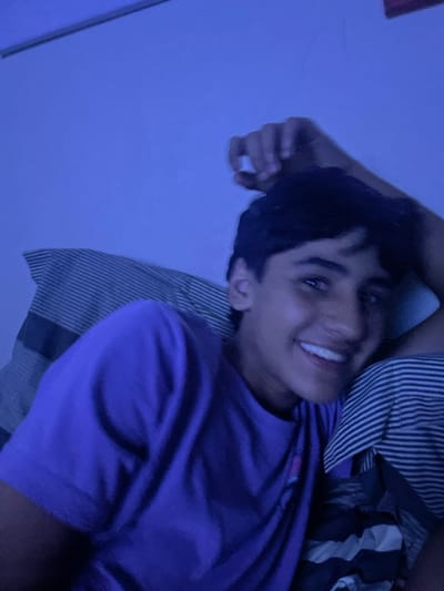 A young man lies on his back on a striped pillow, wearing a bright purple T-shirt. He's smiling, looking directly at the camera. One arm is raised and bent above his head. The image is somewhat dark and appears to be illuminated by a bluish light, giving the scene a cool tone. A striped blanket or comforter is visible near his shoulder.
