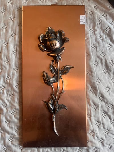 A rectangular, vertically oriented metal plaque, appearing to be copper-toned, is depicted resting on a light-colored, quilted textile surface. The plaque features a three-dimensional relief sculpture of a rose, rendered in a darker, antiqued metallic finish, centrally positioned. The relief displays a large blooming rose at its top, a smaller closed rosebud below it, and a stem with multiple leaves and thorns extending towards the bottom of the plaque. A small white price sticker is adhered to the top right corner of the plaque, displaying "30-10", "QUANTITÉ: 1", and "PRIX: 2,00". The plaque exhibits minor surface scuffs and reflections, with a prominent light glare visible on its right side. The scene is located in Le Puy-en-Velay, France.
