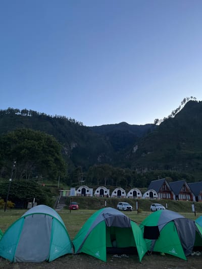 The image depicts an outdoor scene at a campsite in Merodot, Indonesia, likely during dawn or dusk due to the clear, pale blue sky and the subdued lighting. In the foreground, several tents are pitched on a grassy area. Two green and gray tents are most prominent, one open with its interior visible, and another closed. Other tents are partially visible to the left and right.
Behind the tents, a row of distinct dome-shaped structures, possibly cabins or unique camping accommodations, are visible. Further back, several modern wooden houses with dark roofs are situated on a higher level of the terrain. Several cars are parked in front of the houses and dome structures, including two white SUVs and a red sedan.
The background is dominated by a large, densely forested mountain range. The slopes are covered in trees, with some lighter rock formations visible on the cliff faces. The trees at the very top of the mountain range are silhouetted against the sky. The overall environment suggests a peaceful, natural setting, popular for outdoor recreation and lodging.