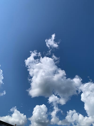The image depicts a clear blue sky with scattered cumulus clouds. In the lower left corner, a dark, angular structure, possibly a fence or railing, is visible. The clouds are white and puffy, with some showing darker grey undersides, suggesting varying densities and thicknesses. The overall scene is natural and serene, characteristic of fair weather. The provided coordinates for Staroe Rakhino, Russia, are not reflected in the image content, as the image solely focuses on the sky and a partial man-made structure at the bottom edge.
