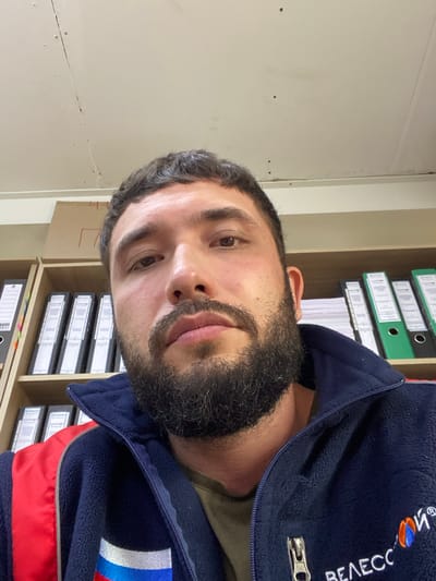 A close-up, selfie-style, medium shot captures a man with dark, short hair and a full beard. He is wearing a dark blue fleece jacket with red accents and a logo on the right side of the chest. Behind him, several shelves of binders are visible, suggesting an office or storage environment. The lighting appears to be artificial, possibly from overhead fixtures, casting a slight glare on the ceiling. The man's expression is neutral. The scene is located in Luzhitsy, Russia.