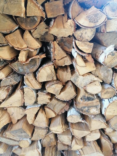 The image shows a close-up, overhead view of a large pile of stacked firewood. The wood is cut into various lengths and thicknesses, with some pieces showing visible bark and grain patterns. The firewood appears to be predominantly birch, indicated by the pale wood and white bark with dark markings. The arrangement of the logs is somewhat haphazard, creating a textured and dense composition. The lighting suggests it is daytime, casting natural shadows across the stacked wood. The location of Lobkovo, Russia, is not visually discernible from the image itself; the scene depicts a common outdoor storage of firewood, likely for domestic heating. There are no people or actions depicted in the image.