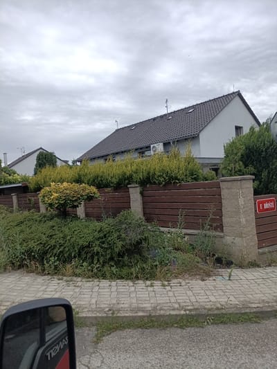 This is a photograph taken from a vehicle, showing a residential area on a cloudy day. In the foreground, the side mirror of the vehicle is visible, reflecting a portion of the vehicle's interior and a red and white logo with the text "TIEMAS" in reverse.
Beyond the vehicle, a paved sidewalk runs alongside a wooden fence. The fence is constructed with horizontal dark brown wooden planks supported by concrete pillars. A small, ornamental tree with yellow and green foliage stands in front of the fence, surrounded by lush green bushes. Behind the fence, two houses are visible. The house on the right has a dark grey tiled roof and a light grey exterior wall. An outdoor air conditioning unit is mounted on its wall. The house on the left also has a grey tiled roof and a white exterior.
Attached to one of the concrete fence pillars is a red sign with white text that reads "U HŘIŠTĚ". This translates to "At the playground" in Czech, suggesting the location of the property. The overall atmosphere is overcast and still, with no people visible in the frame. The scene depicts a quiet suburban or semi-rural setting in Svémyslice, Czechia, likely during the daytime.
