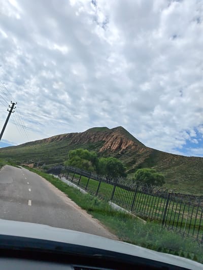 A paved road with white dashed lines curves to the left, leading towards a verdant, hilly landscape. A dark metal fence runs along the right side of the road, bordering a grassy area and scattered trees. Beyond the fence, the hillside is covered in green vegetation, with exposed reddish-brown rock formations visible on the peak of a prominent mountain. Power lines and a utility pole are situated on the left side of the road, ascending towards the sky. The sky is filled with a mix of white and gray clouds, with patches of blue visible. The image appears to be taken from a vehicle, with the top of the car's dashboard visible in the foreground. This scene is consistent with the natural beauty of the mountainous region surrounding Cholpon-Ata, Kyrgyzstan, likely depicting a road leading into or out of the city towards the Issyk-Kul lake or the surrounding terrain.