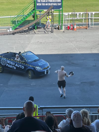 In Punchestown Racecourse, Ireland, on a paved surface, a dark blue convertible car with the text "IRISH AUTO PARTS" and "Timeless Marketing" visible on its sides, drives towards the right side of the frame. The car has yellow accents on its wheels and along the top edge. Inside the car, a person wearing a light-colored balaclava and a tan shirt is seated in the driver's seat, holding a steering wheel. Another person, wearing a tan full-body suit and black shorts, is on the pavement to the right of the car, holding a helmet in their right hand and facing away from the car. The background features a grassy area, a green metal staircase leading to a ramp, and various barriers and cones. In the foreground, spectators are seated in bleachers, with their backs to the camera, observing the scene.