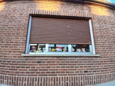 The image shows a brick building with a window that has a brown rolling shutter. Inside the window, on the ledge, are several stuffed animals and figurines. The stuffed animals include what appear to be teddy bears, a figure with a red hat, and other animal figures. The figurines include a Santa Claus figure, a yellow character, and what looks like a cow and a rabbit. The window is set into a red brick wall. The image was taken in Avila, Spain.