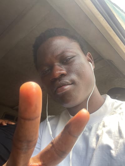 A close-up, front-facing image shows a young individual, appearing male, with dark skin and short dark hair, posing within an enclosed space in the city of Aiyetoto-Asogun, Nigeria. The subject wears a white short-sleeved crew-neck t-shirt and white wired earbuds are inserted into both ears, with the wire running down the front of the shirt. The individual looks directly into the camera, with lips slightly parted and a slight sheen visible on the facial skin. The subject's right hand is raised into the foreground, extending the index and middle fingers to form a 'V' shape. In the upper right background, the top of another dark-skinned individual's head is partially visible. A dark-colored garment, possibly belonging to a third person, is partially visible in the lower left corner. The immediate background suggests the interior of a vehicle, possibly a car or bus, with light-colored ceiling panels and a vertical structure, likely a window or door frame, visible on the right side.