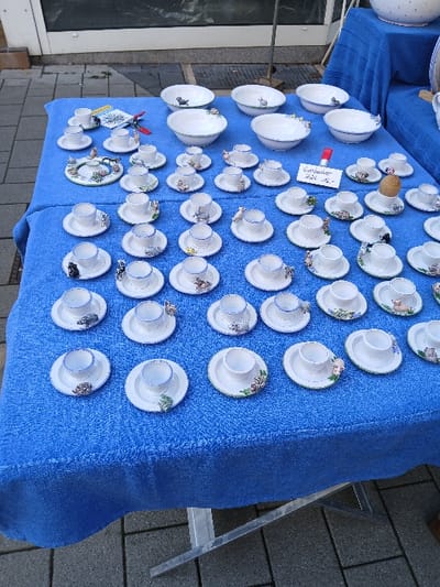 The scene depicts an outdoor market stall in Homburg, Germany. A rectangular table is covered with a textured, bright blue fabric. On this table, a large collection of white ceramic dinnerware and novelty items are meticulously arranged for display.

The foreground and midground feature numerous small, white ceramic egg cups, estimated to be over 50 individual pieces. Each egg cup possesses a blue rim and is accompanied by a small, integrated saucer or base, often adorned with a unique, miniature hand-painted figure, including various animals such as pigs, cats, and birds, as well as floral motifs. These small items are arranged in organized rows and clusters. A small white sign with red text, partially visible, indicates "Eierbecher...Stück 6,-," indicating "egg cups...6 each."

Towards the rear of the table, approximately six to eight larger, white ceramic bowls are displayed. These bowls feature a green rim and contain similar small, painted animal figures, such as rabbits, pigs, and cats, or floral designs on their interior surfaces.

To the right, a second table, also draped in blue fabric, holds additional ceramic items, including a large white bowl with blue polka dots, partially visible. The immediate background shows a building facade with a large reflective window or door. The ground surface beneath the table consists of grey interlocking pavers or cobblestones. The scene is illuminated by bright, diffused daylight. No individuals are present, and no active interactions are depicted.