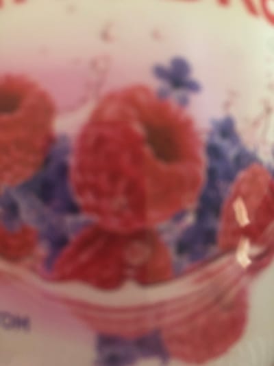 The image is a blurred, close-up shot of a product label, likely for food items. Dominating the foreground are abstract shapes in red and pink, suggesting fruits such as raspberries, possibly arranged in a pattern or forming part of an illustration. Interspersed with these are out-of-focus blue elements, which could represent flowers or other decorative motifs. In the lower left corner, a portion of text is visible, with the letters "OM" being partially discernible. The overall impression is one of a stylized and colorful design, with no specific indication of location in Luka-Meleshkivska, Ukraine.
