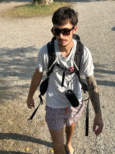 A young man with dark hair and a goatee is captured in a medium, high-angle shot, walking outdoors. He is wearing a white t-shirt, patterned swim trunks, and sunglasses. A black backpack with straps is visible on his back, and a black cylindrical object, possibly a portable speaker, is attached to the backpack. His left arm, tattooed with intricate black designs, is partially visible. The man appears to be walking on a gravel path, with patches of green grass to his left and shadows cast by trees on the ground to his right, suggesting a sunny day. The image is taken in portrait orientation, with the subject positioned slightly off-center to the left. There is a small red and white tag visible on the chest strap of his backpack. The overall impression is of a casual, outdoor excursion.