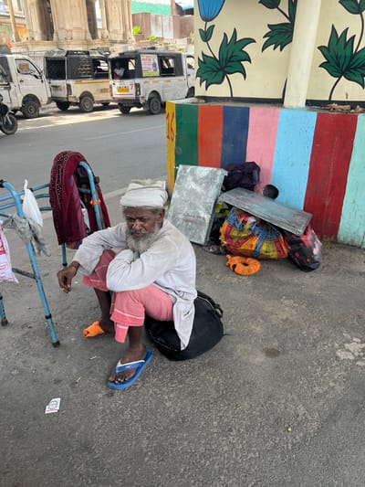 An elderly man with a white turban and a long gray beard sits on a black bag on the side of a street in Ajmer, India. He wears a light-colored long-sleeved shirt and pink pants. His right hand is raised and he appears to be holding a cigarette. He is wearing blue flip-flops. To his left, a blue walker with a red garment draped over it stands near a white plastic bag. Behind him, a collection of belongings including bags and what appear to be metal sheets are piled against a brightly colored wall with a painted floral design. In the background, several white vehicles, possibly auto-rickshaws, are parked on the street. The ground is asphalt.
