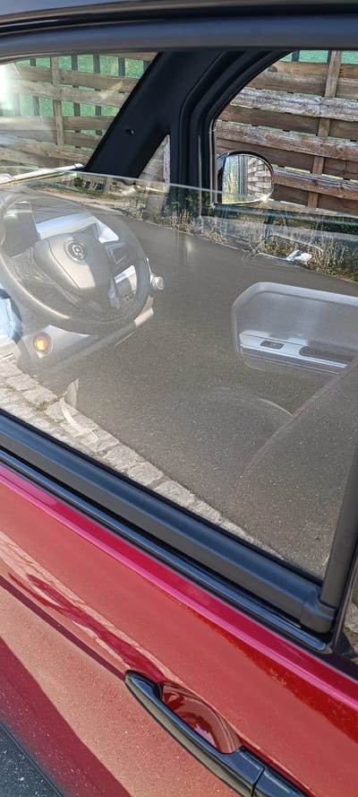This media file captures an interior view of a red car, taken from the driver's perspective. The scene is oriented vertically, suggesting the photo was taken with the phone turned on its side. The foreground shows a portion of the steering wheel with a circular logo, and a glimpse of the dashboard with an orange indicator light. A portion of the car door frame, with its black rubber seal, is also visible on the left.
Reflected in the car's window is an outdoor scene. We see a wooden fence with vertical planks, which appears to be a location cue for Mühlhausen, Germany. Sunlight glints off the surfaces, indicating it is daytime, likely sunny. The reflection also shows a small side mirror on the car, reflecting more of the fence and sky. The car itself is red, with a black door handle visible on the right side. The ground outside appears to be asphalt or a paved surface. There is no discernible text within the frame or its reflections. The overall impression is a mundane moment, possibly a pause in driving or a quick photo opportunity.