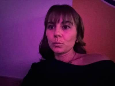 A woman with bangs stares directly at the viewer. Her face is illuminated with a pinkish-red light. She wears a dark top. The background behind her head is divided into two areas; one is a vibrant orange color, and the other is a shade of purple. The image is rotated at 180 degrees. She has an earring on her left ear and is wearing a necklace.
