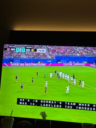 The image shows a television screen displaying a soccer match. The score is NOR 3, SEN 1, at 79:08 minutes. The match is part of the FIFA World Cup 2022, Group 1. The field is green, with players in black and white uniforms. A banner across the bottom of the screen reads "OVER 160 DESTINATIONS" and "QATAR AIRWAYS". Another black banner at the bottom of the screen displays text that appears to be a caption: "HE'S IN NORWAY'S TEAM WHEN H WAS 15, LABELED THE TWONDER". In the foreground, out of focus, are a white paper cup with a straw, a dark baseball cap with an orange logo, and a crumpled white tissue. The television is positioned in what appears to be a living room or entertainment area, suggested by the dimly lit background and the presence of personal items. The overall scene suggests a casual viewing of a sporting event. There is no visible indication that this scene is located in the city of North Bergen, United States of America.