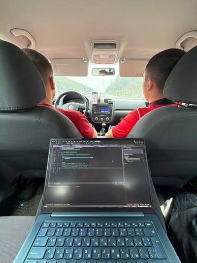 This image captures the interior of a moving vehicle from a rear passenger's perspective, looking forward. Two males occupy the front seats: a driver on the left and a passenger on the right. Both individuals have short dark hair and are wearing red short-sleeved shirts, the passenger's shirt featuring a black collar detail. The vehicle is a Volkswagen, evidenced by the emblem on the steering wheel. A smartphone displaying a navigation application with a highlighted route is mounted centrally on the dashboard. Below the phone, the car's audio/infotainment system and climate controls are visible, along with a manual gear shift lever. In the immediate foreground, an open, dark-colored laptop rests on a lap, its screen displaying lines of code within a dark-themed integrated development environment. The laptop's keyboard features both Latin and Cyrillic characters. Through the front windshield, a multi-lane road is visible, bordered on both sides by forested hills under an overcast sky. The scene is situated on a road in the vicinity of Церово, Bulgaria.