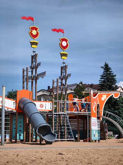 The image depicts a children's playground designed to resemble a pirate ship, located in the city of Myadzyel, Belarus. The playground features a large, orange and gray slide, rope nets, and wooden structures that mimic a ship's masts and deck. Two young boys, wearing swim trunks, are visible on the upper deck of the playground structure, appearing to interact. A young girl in a dark outfit stands near a ramped entrance to the playground. The playground is situated on a sandy surface, with residential buildings and dense evergreen trees visible in the background under a partly cloudy sky. Two prominent flags with red banners and decorative shields are mounted on tall poles, adding to the pirate ship theme.