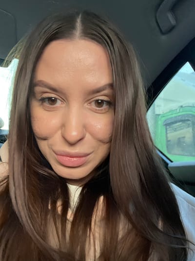 A female subject with long, dark brown hair parted in the middle is positioned centrally, looking directly at the camera with a slight smile. She has brown eyes and appears to be wearing a light-colored top. The setting is identified as the city of Svinoedovo, Russia. The background suggests she is inside a vehicle, with a dark headliner and part of a car pillar visible on the right. Through the vehicle's window on the right, a blurry, predominantly green structure or object is visible in the exterior environment. The overall lighting is soft and even.